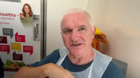 Ian Stewart, sitting in the mobile testing van wearing a translucent medical apron and blue shirt, looking slightly out of frame.