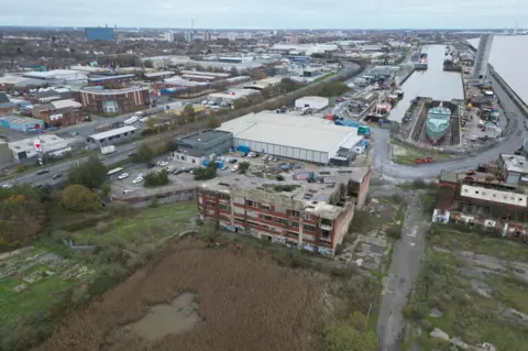 A drone view of docklands and a riverside, with the buildings of Hull behind them. In the foreground, an old dock is partially filled in. It is covered in reeds with patches of water. Old dock buildings can be seen around it. In the middle distance, ships can be seen in another dock.