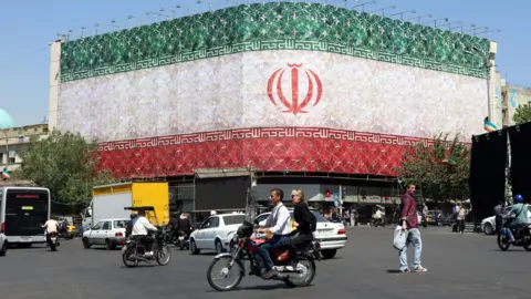 Iranians drive cars and motorbikes past a billboard featuring Iran's national flag at Enqelab Square in Tehran, Iran.