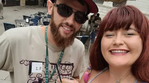 Chelsea Rees holding a drink. The mole is visible on her chest jus above her top. She is stood next to a man who has a beard and a beige t shirt on.