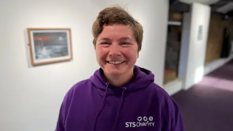 Catherine Scott-Jones, wearing a purple hoodie with a white logo saying 'STS Charity', smiles at the camera. She is standing in front of one of the works created by David Hughes. 