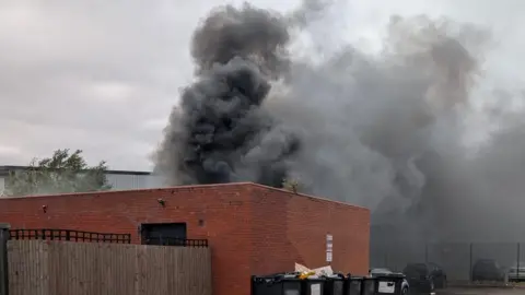 Supplied A fire in a building, showing thick dark smoke coming up in the air. There is a row of black bins, cars in the distance and a building behind the smoke. 