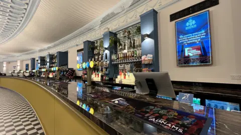 Shaun Whitmore/BBC A view of a long bar, showing beer pumps, optics, and a beer mat in the foreground with Adnams written on it. The floor is a black and white check.