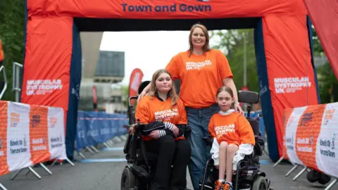 Muscular Dystrophy UK/Chris O’Donovan The Blackmore family next to the race start-line. The mother is stood up with her arms around her two daughters - who are both in wheelchairs.