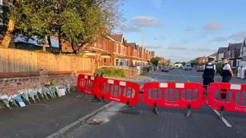 Road closed off with a red barrier. Floral tributes are left against a nearby wall and two policeman are stood at a cordon 