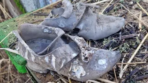 Chris Fellows A pair of old shoes placed on a wooden table covered with thatch straws. They are discoloured and worn off. 