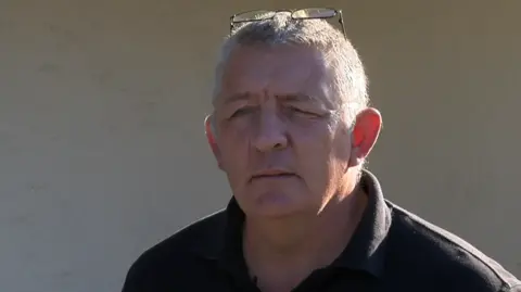 Paul Bourgaize. He has short grey hair and a pair of glasses on his head. He is wearing a black t shirt. 