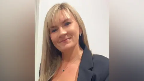 Carol Georgiou stood in front of a white wall, wearing a black suit jacket and a silver necklace. She has long blonde hair and is smiling.