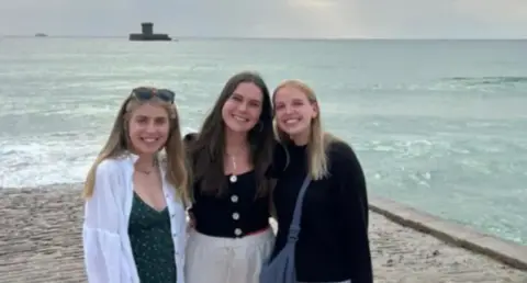 Ella Jennings Three smiling women stand in front of a beach and coastline looking at the camera