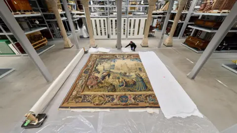 David Levene/Art Fund/PA Media A huge wall hanging is rolled out across the floor, with a cloth on one side and polythene coverings underneath. A man is kneeling nearby. There are ancient-looking columns behind him, with items on warehouse shelves around him.