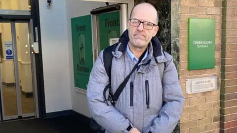 Local Democracy Reporting Service A man with glasses and wearing a grey coat is standing outside a branch. A door is on the far left and a green sign with the words Tower Square Tunstall in white is on the right.