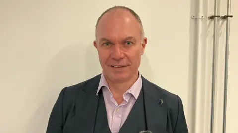 A man in a grey suit , lilac shirt and stood in front of a white wall 