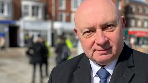 Carl Smith is standing in a market place in Great Yarmouth. He is looking directly at the camera and is wearing a white shirt, navy blue tie and a black coat.