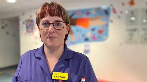 A woman in a nurses uniform is looking at the camera. She has red hair and red framed glasses. She is wearing a yellow name badge with "Charlotte" written on it. 
