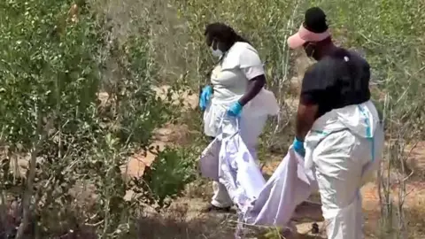 Reuters Two detectives in mainly white protective clothing (one is wearing a black T-shirt) and facemasks carry a white body bag through bushes with the exhumed remains of one of the suspected victims of a religious cult buried in a shallow grave in Kwa Binzaro village - 21 August 2025