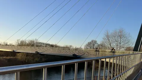 Worcestershire County Council A closer shot of the above bridge, showing the bridge as it curves over the river. Several people are walking across it.