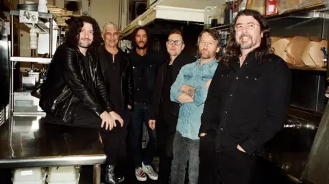 Elizabeth Miranda A promotional photograph of the Foo Fighters, showing the band in the catering area of a music venue, with bulbs of garlic, cooking oil and takeaway boxes in the background. Drummer Ilan Rubin is perched on a table, while the rest of the band stand in a semi-circle, looking into the camera.