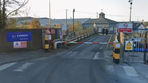 The entrance to the 2 Sisters Food Group in Willand. There is a red and white barrier over a lane and signage to the left reading 2 Sisters Food Group and a warning to pedestrians. To the right is a sign which reads: No Sounding Horns and a second one which reads: Caution Automatic Barrier. There are two people at the end of the short lane walking towards a single storey factory building.