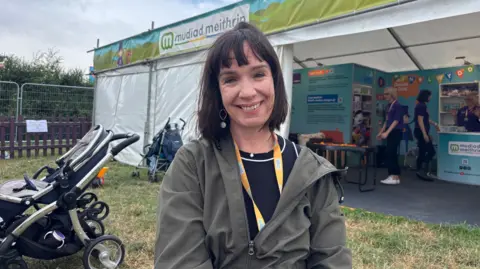 Gwenllian Landsown, chief executive of Mudiad Meithrin stands outside their promotional stand at the Eisteddfod. She wears a khaki jacket, has a brown bob and smiles at the camera.