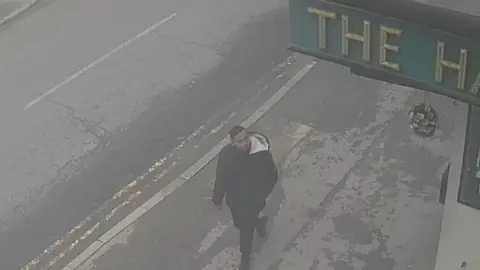 West Yorkshire Police A CCTV image in black and white showing a man with a close-cropped beard and short hair walking along the pavement beside a main road.