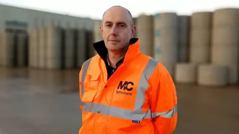 A bald man in a high-viz orange FPMcCann branded jacket, stood at an industrial plant with concrete pipes visible behind him