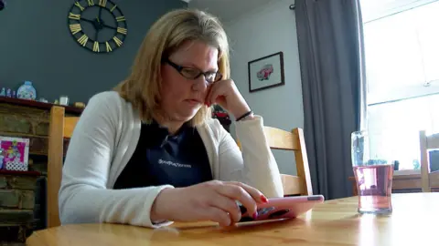 Samantha O'Sullivan sitting at a wooden table with her phone in her hand. She is watching the screen. There is a glass on the table, to her right, and a clock on the wall behind her. She is wearing a blue top with a white cardigan.