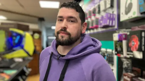 Louis Phelps stands in his store wearing a purple hooded top. He faces the camera with a serious expression on his face.  