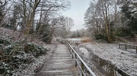 BBC Weather Watchers/Stormchaser Al A frosty wooden bridge is surrounded by trees and frozen water. In the distance, a lake is frozen with more trees around the edges. 