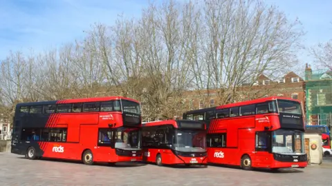 Three red electric buses lined up in front of buildings in Salisbury. 