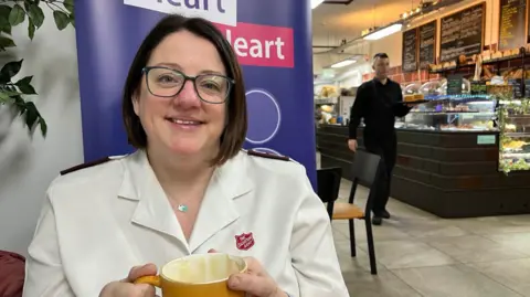 Major Alison Friday in The Salvation Army uniform holding a cup of coffee with The Canterbury Bakery waiter in the background