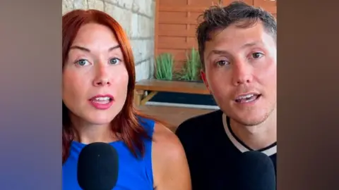 Bec and Sam sit side by side indoors speaking into black microphones. A concrete block wall, wooden panelled screen and potted plants are visible behind them.