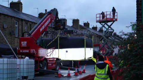 Image shows a closed street with a crane and lighting on it. Cones are on the street and a cherry picker has camera crews on it.
