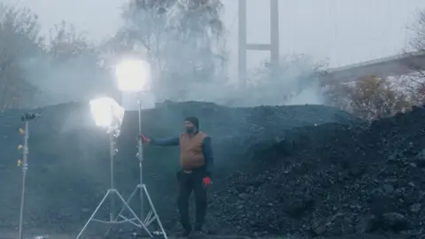 Northern Films A crew member stands with two film lights in front of a mound of rubble with the Humber Bridge and trees in the background.
