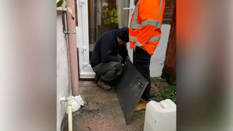 A man crouches down in a back yard, next to a door to a home, and works on a flood door on the ground. A person wearing an orange hi-vis jacket is standing next to him.