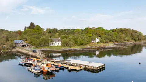 Sothebys International Realty The Boat House and whitewashed cottages sit beyond the jetty, where several boats of various sizes are moored.