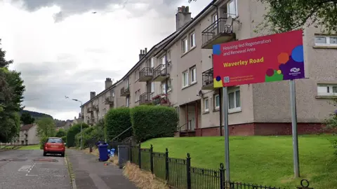 Residential street with low-rise apartment blocks and a sign reading 'Housing-led Regeneration and Renewal Area – Waverley Road', grassy verge and parked car