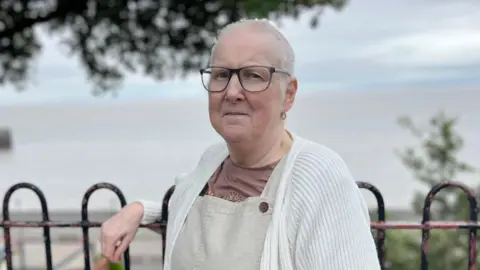 Sally is leaning on railings with the sea behind her. She is wearing a cream cardigan, brown t-shirt and light brown dungarees. She has short white hair and glasses.