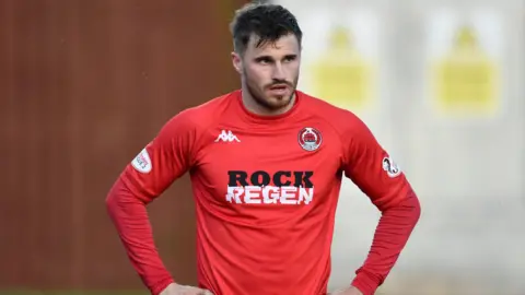 SNS Footballer David Goodwillie stands with his hands on his hips, while wearing a red long-sleeved football top.