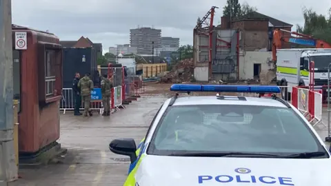 A police car is parked in the entrance of an industrial site. Military personnel talk to each other further back.