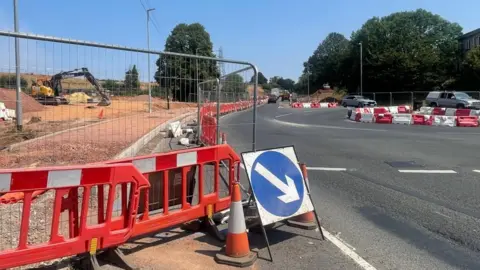 Roadworks at a junction with fencing, cones and road signs and a digger in the background.