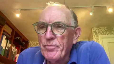 Visit Scilly chairman Andrew Sells. He is an elderly man with short grey hair. He is wearing round green-rimmed classes and a blue polo shirt. He is sat in a room with shelves holding a number of folders. The room has floral wallpaper.