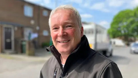 Nicola Rees/BBC A man with short grey hair smiling at the camera, wearing a black jacket with a large collar