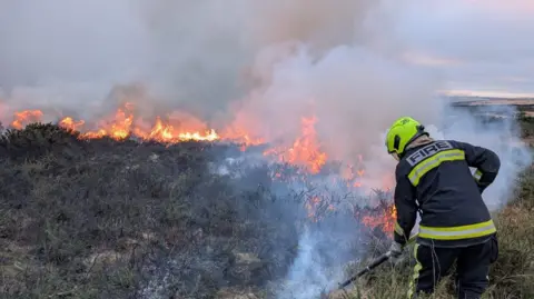 Devon & Somerset Fire & Rescue Service The image shows a firefighter in protective clothing standing at the edge of a moorland wildfire and using a swatter to try and extinguish the flames. Gorse and vegetation is ablaze in the background. 