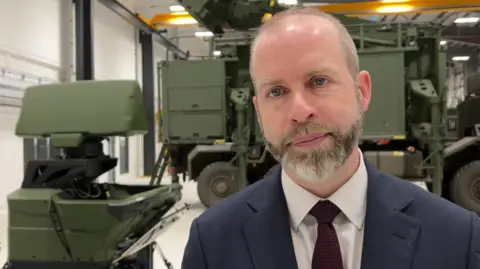 Jonathan Reynolds, wearing a dark blue suit jacket, white shirt and maroon tie, stood in front of a military vehicle in a factory.