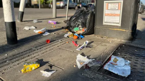 Litter in Victoria Park, Cardiff. There is a black bin bag which is split and there are crisp packets, papers, and fruit strewn across the floor.