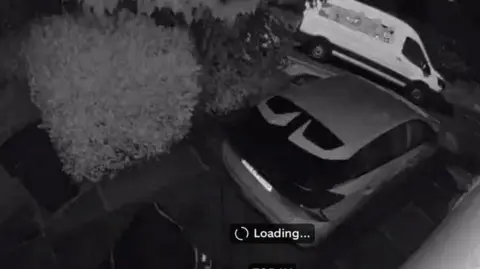 Mandy Greenwood A black-and-white night-vision CCTV still showing a driveway. In the foreground, there is a parked car. Behind the car, a large white van is parked on the street. 