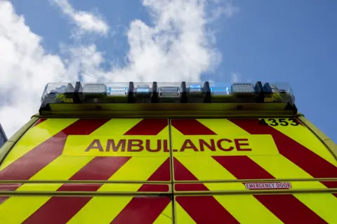 A generic picture of the back of an ambulance. The vehicle has the word 'ambulance' on it and has red and yellow markings and the lights on top. 