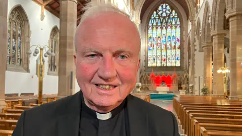 Bishop Donal McKeown is standing inside a cathedral, wearing a black jacket and shirt with white clerical collar. Rows of church pews and an altar are in the background