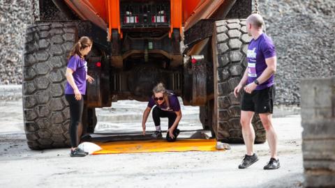 A woman crawls under a large digger in Ronez Quarry while another woman checks by to see how see is doing and a man is about to run on in the True Grit challenge