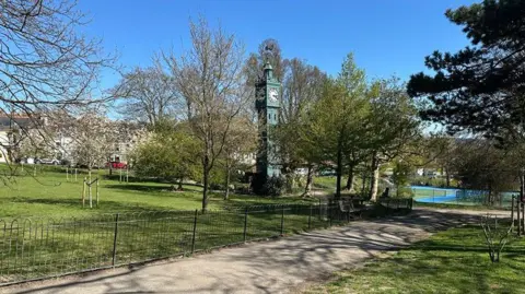 Blakers park in Brighton has a wrought‑iron fence and green lawn leading towards a tall ornamental clock tower, surrounded by trees, with a tennis court visible in the background under a clear blue sky.